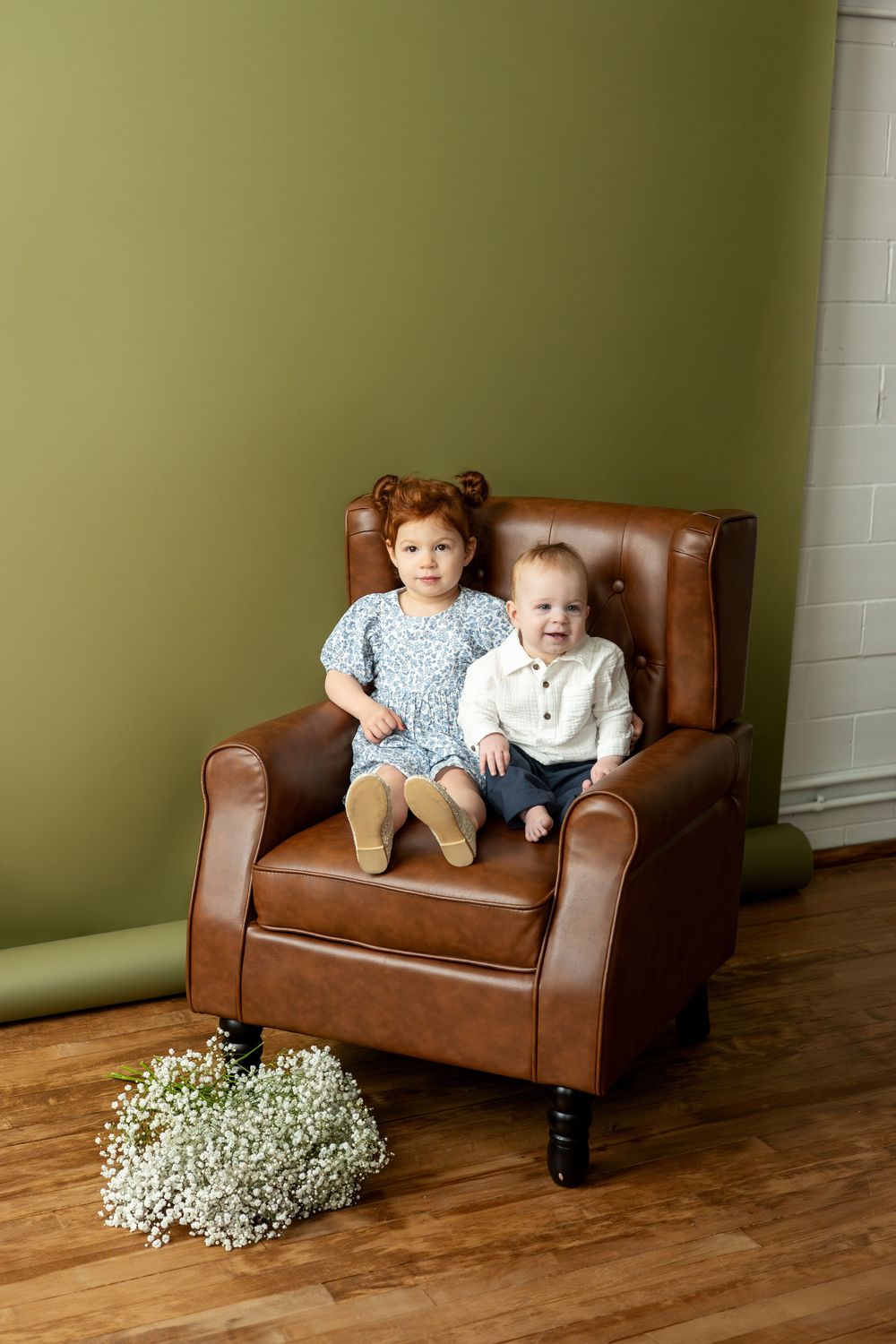 Two young siblings sitting on brown tufted leather chair with white baby's breath flowers on floor.