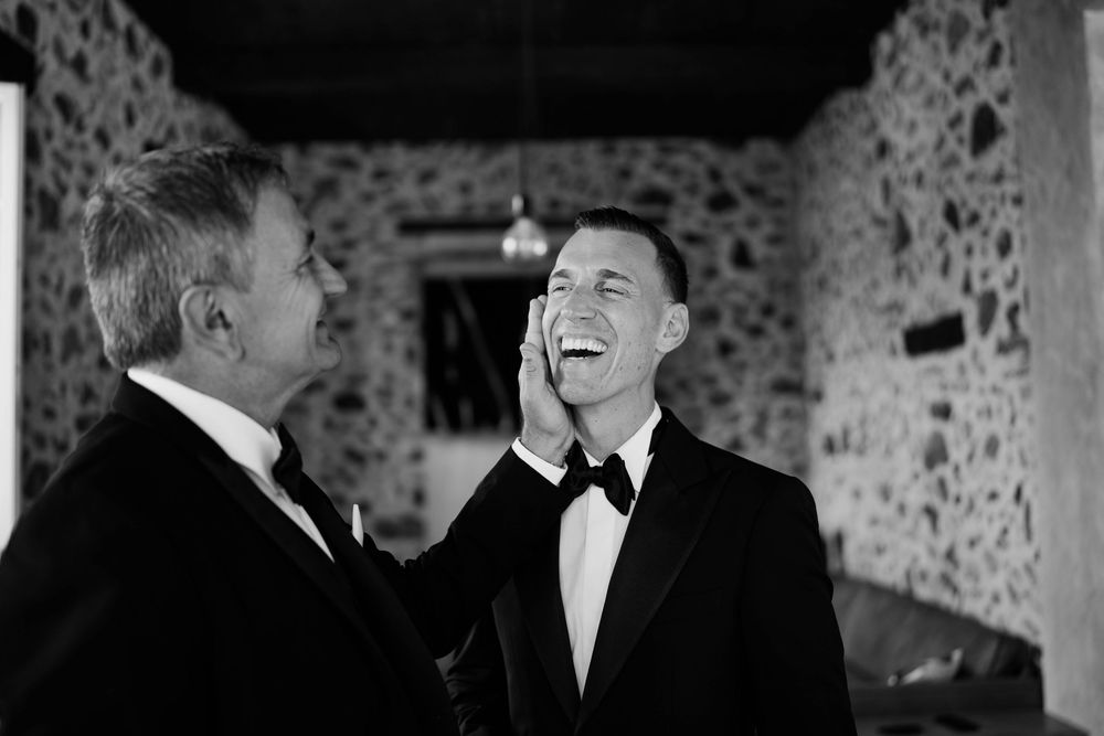 A candid black and white moment of joyful laughter between two men in tuxedos at an elegant stone-walled venue.