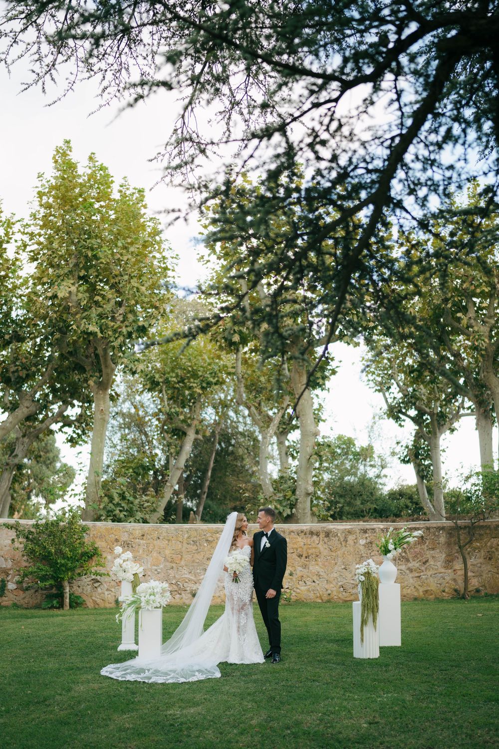 Couple de mariage et fête de mariage en tenue formelle posent ensemble sur une pelouse herbeuse contre une toile de fond de mur de pierre.