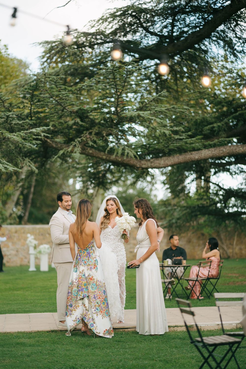 Fête de mariage en robes à fleurs et blanches se rassemble sur une pelouse avec des guirlandes lumineuses et des arbres au-dessus au coucher
