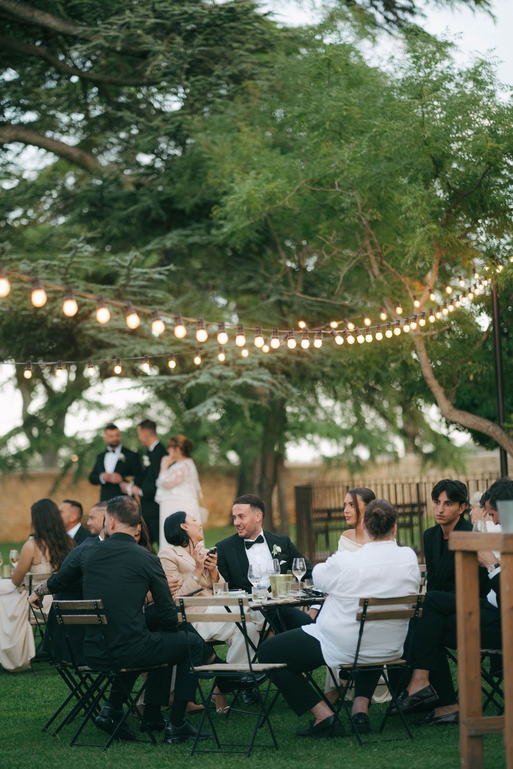 String lights illuminate an outdoor evening wedding reception with guests seated at tables under tall trees.