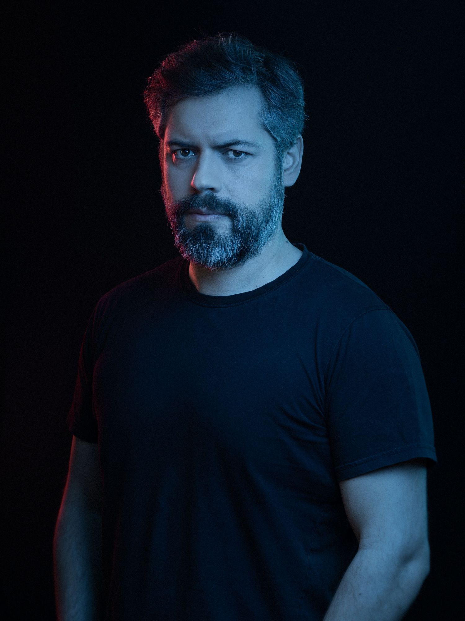 Bearded man in black t-shirt poses against dark background with dramatic blue and red lighting.