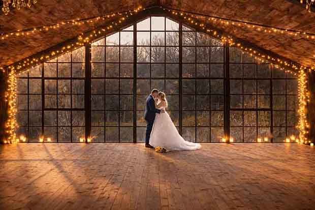 Bride and groom embracing in a rustic barn venue with warm string lights and large windows at sunset.