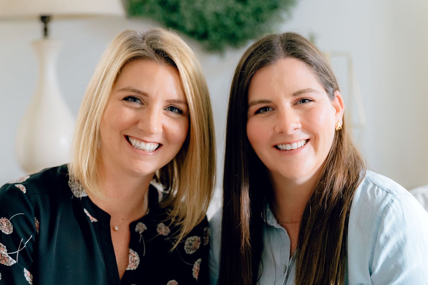 Two women with bright smiles sit together wearing black and light blue tops in a casual indoor brand photo session in Hudson, Ohio home.