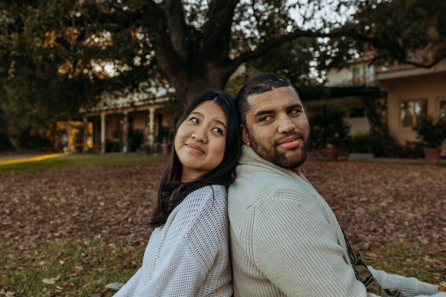 A couple sits back-to-back outdoors in autumn, wearing cozy sweaters while smiling on fallen leaves at Will Rogers State Park.