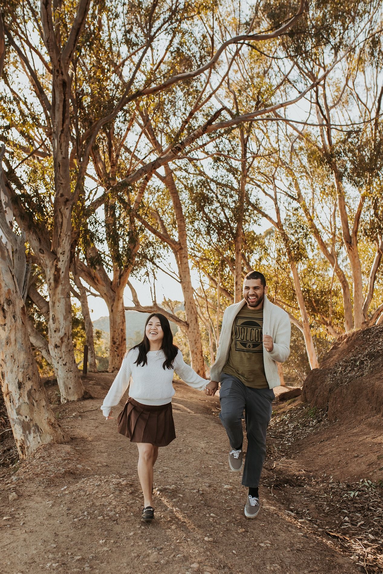 A couple walks along a dirt path surrounded by tall eucalyptus trees in warm sunset lighting at Will Rogers State Park.