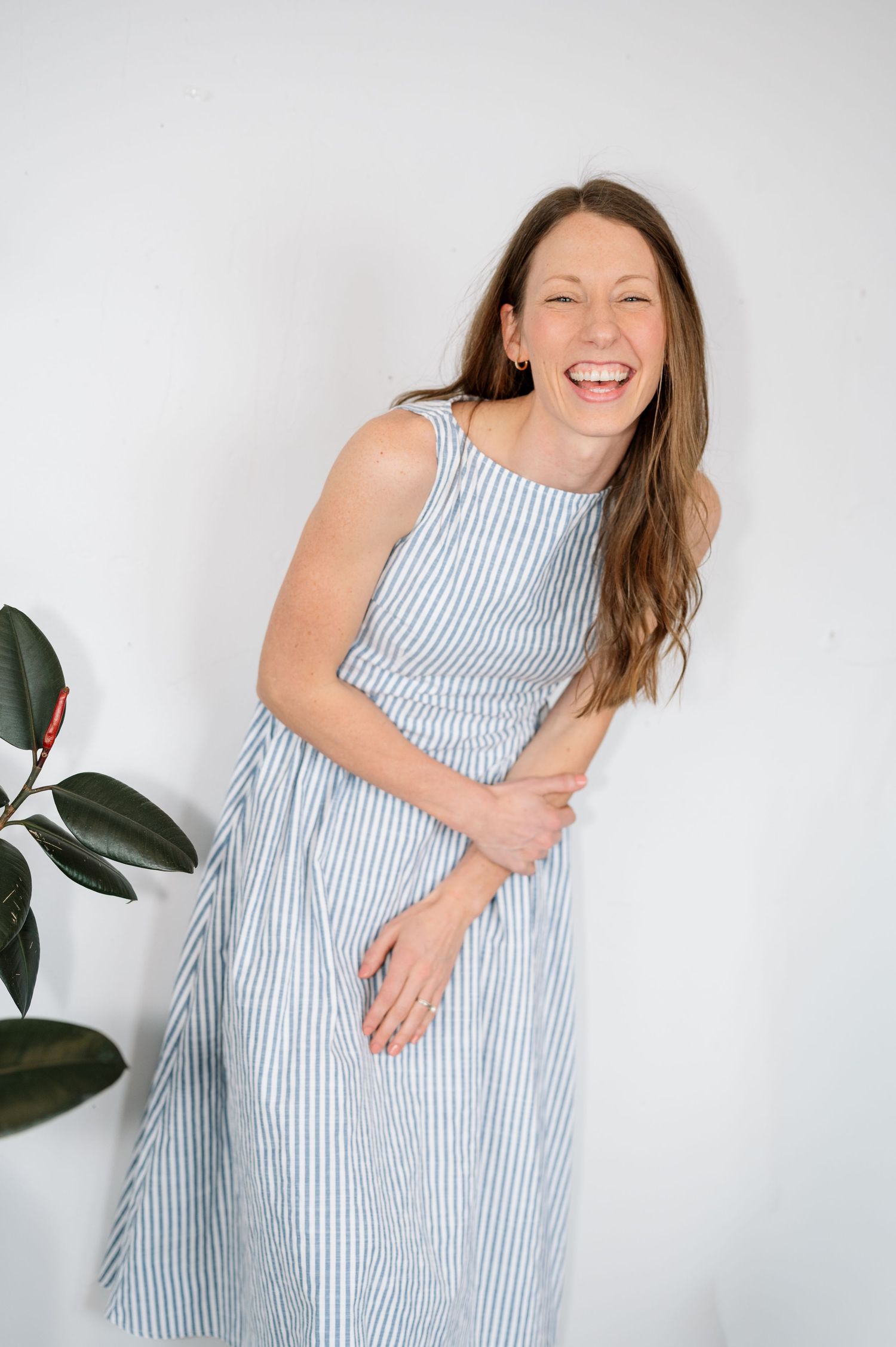 A person wearing a blue and white striped sleeveless dress laughs joyfully against a white wall with a plant nearby.