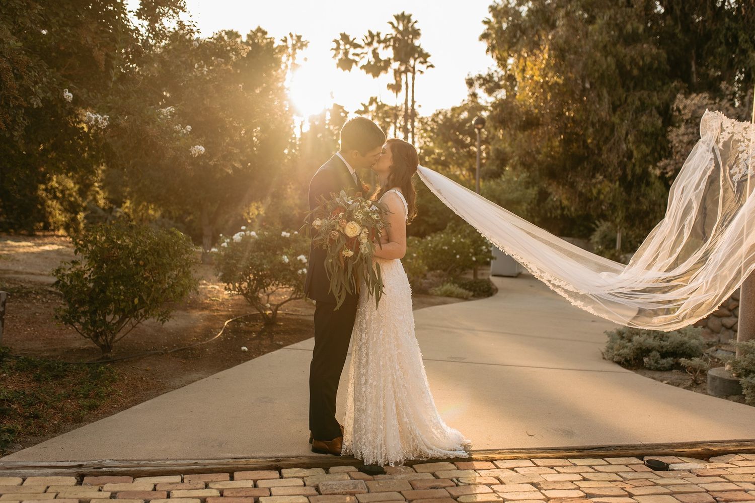 Bride and groom share a kiss at golden hour, with veil flowing in the breeze amid lush trees at Rancho Buena Vista Adobe.