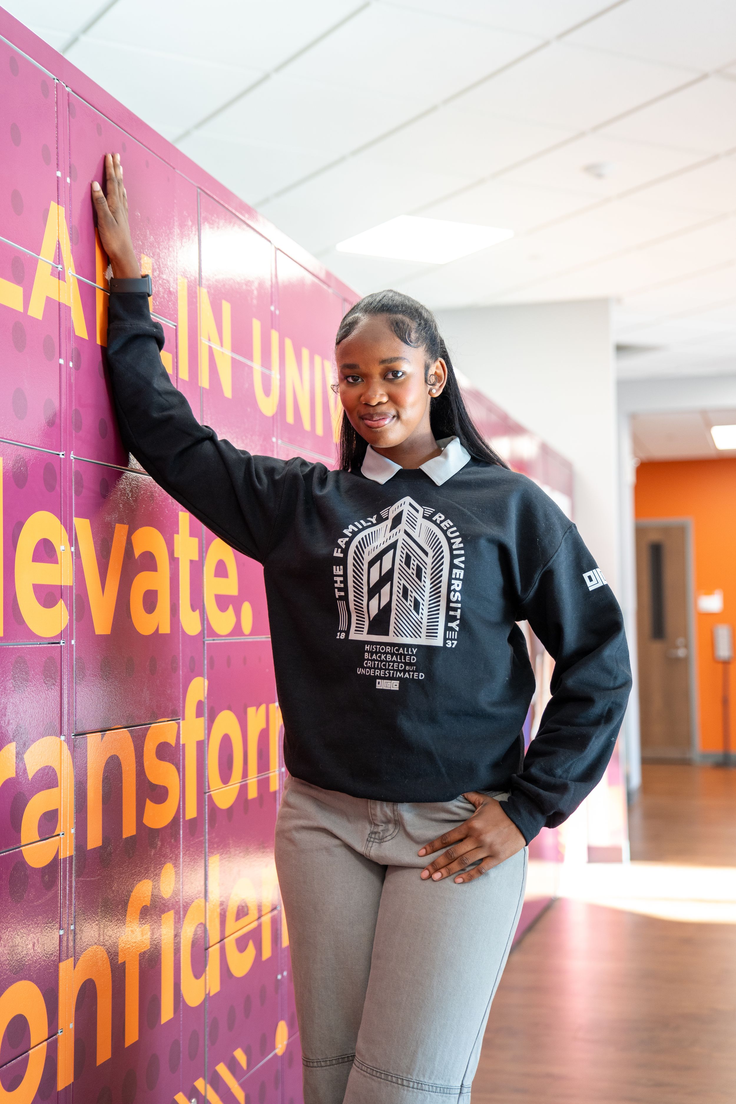 Smiling student in a dark university sweatshirt leans against a purple wall with yellow lettering inside a Clafin University building.