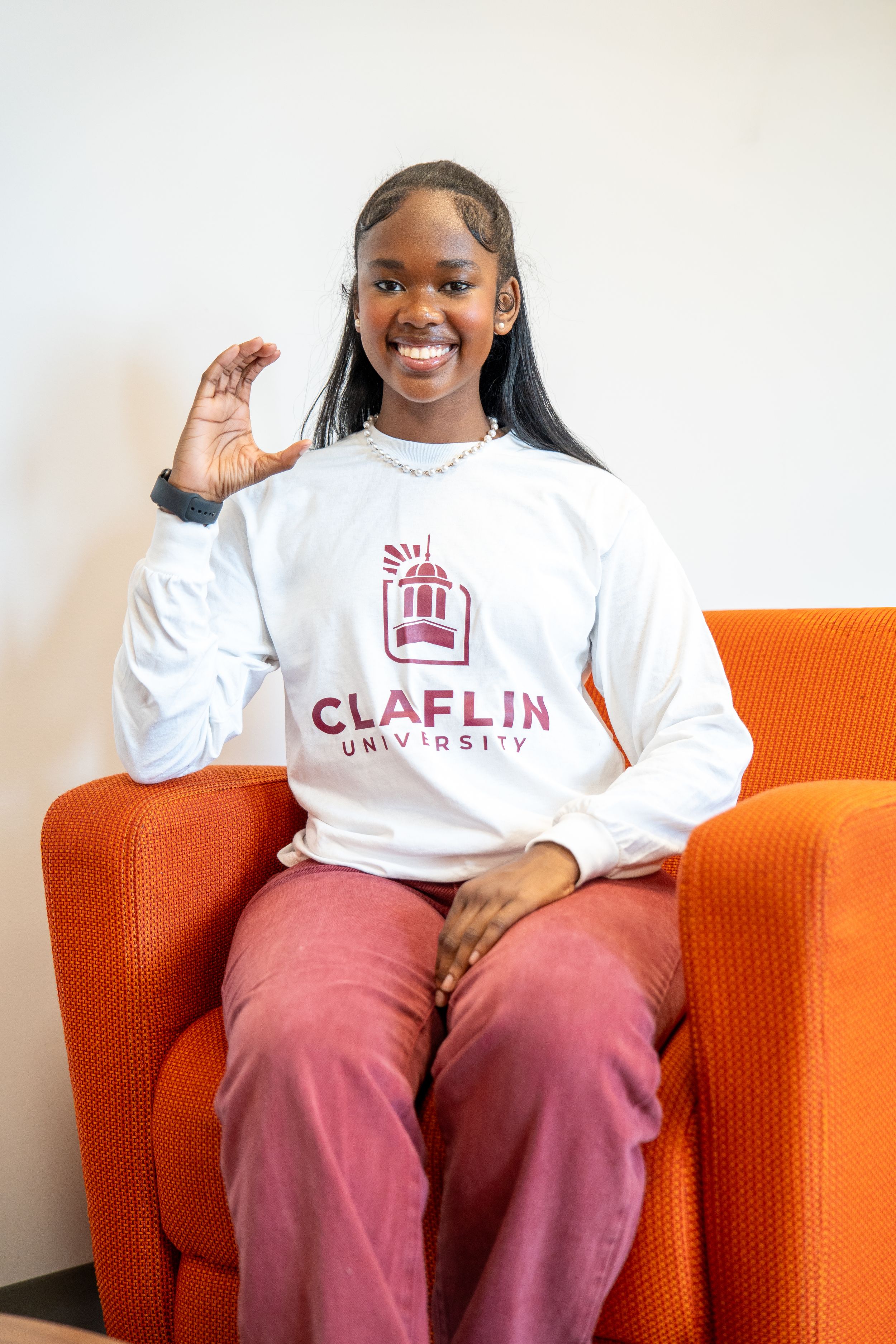 Smiling student in a white Claflin University sweatshirt and pink corduroy pants sits in an orange chair making a hand gesture.