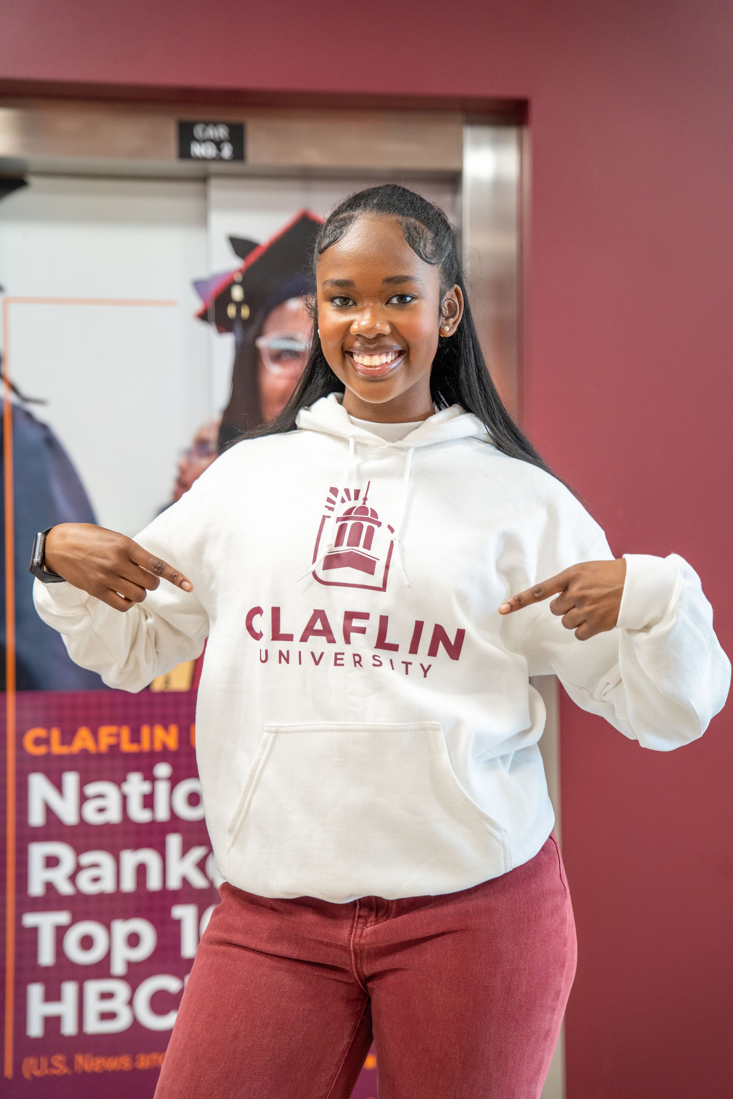 A smiling student in a white Claflin University hoodie points to herself in front of a poster about a top HBCU ranking.