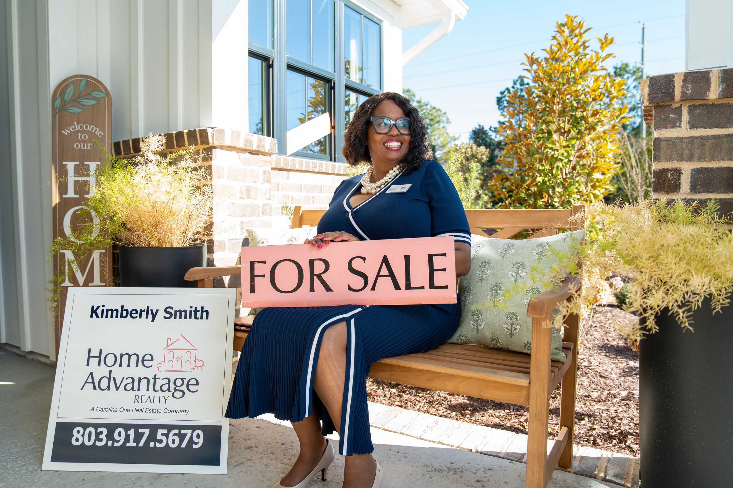A woman in a blue dress sits on a porch bench holding a For Sale sign beside a Home Advantage Realty yard sign.