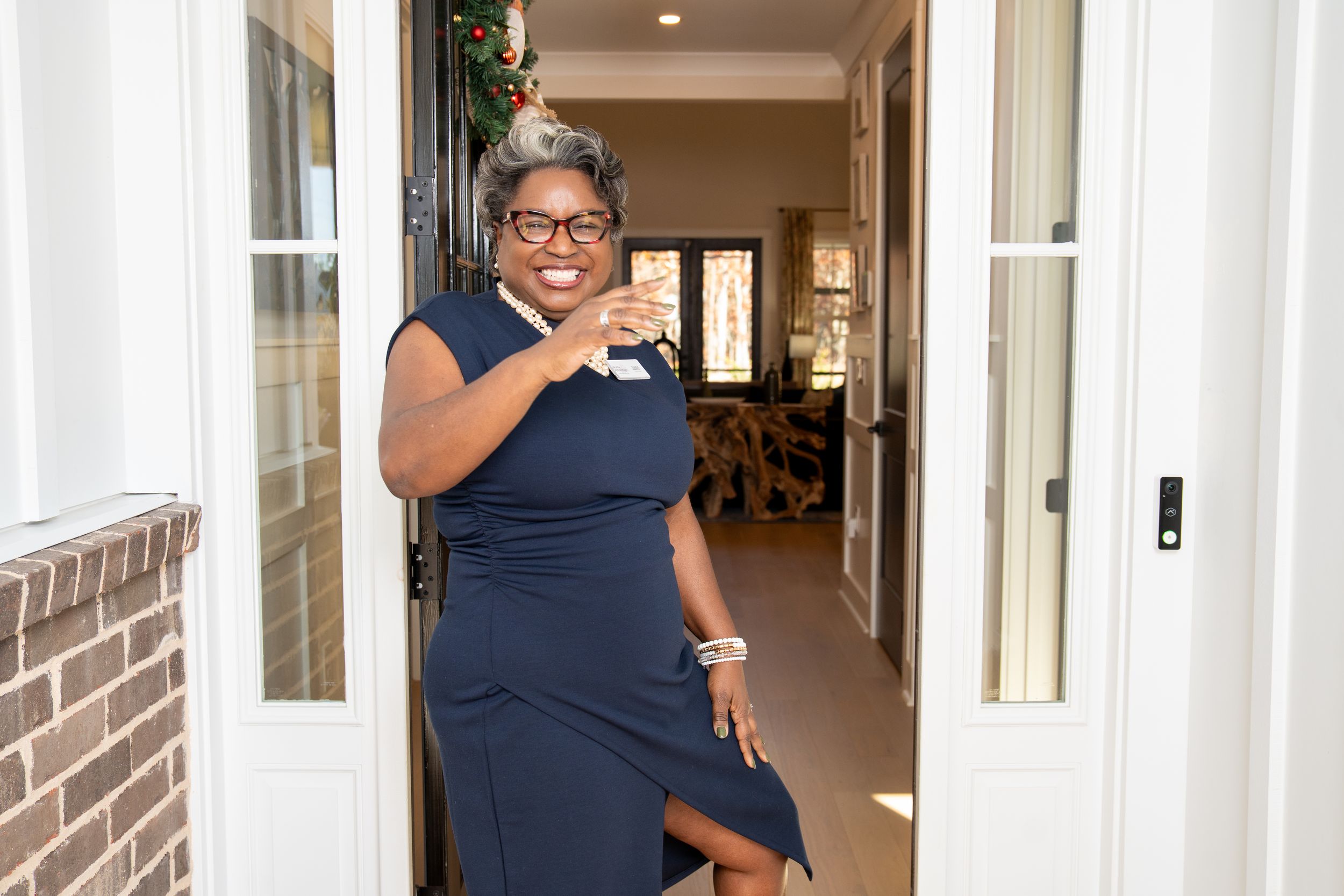 A smiling real estate agent in glasses and a navy dress laughs in a bright doorway of a brick home.