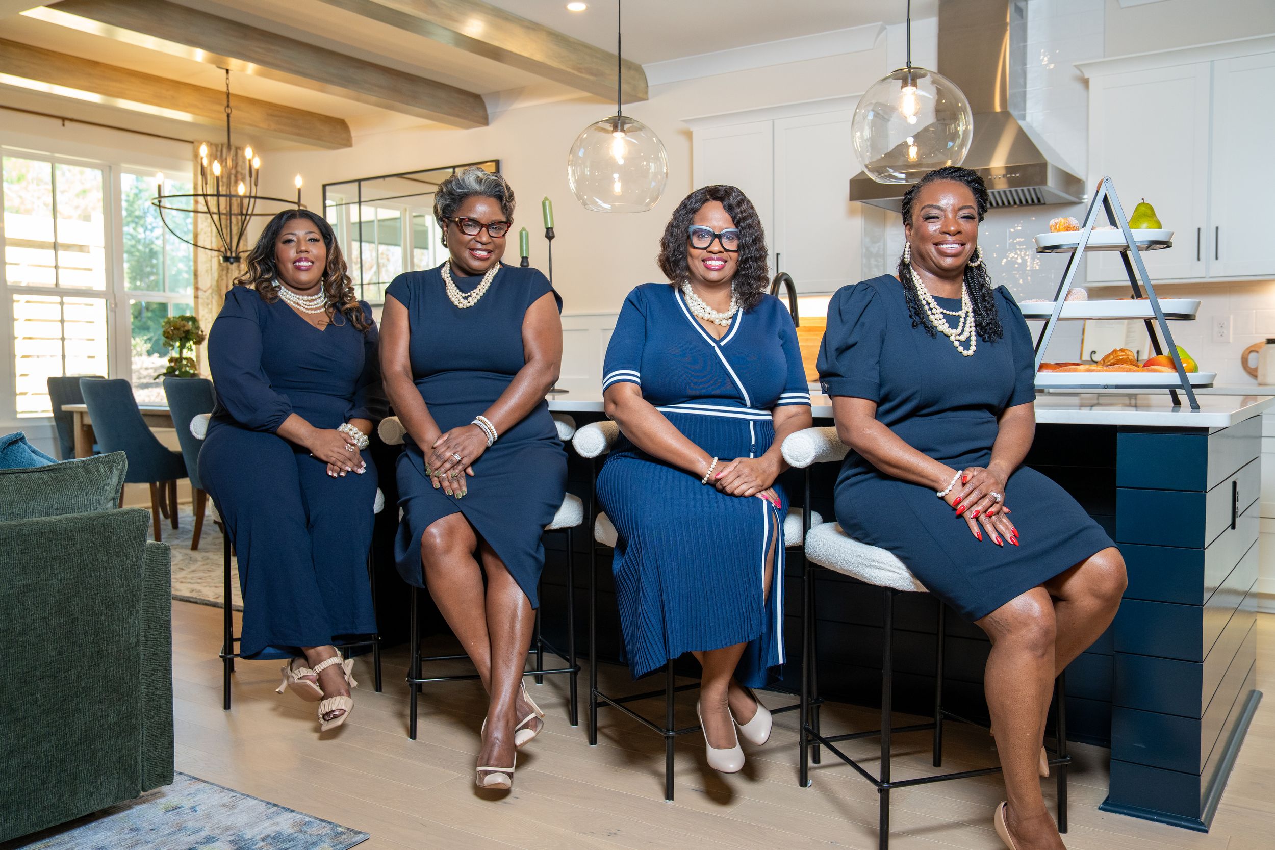 Four women in matching navy blue dresses and pearl necklaces pose together in a modern kitchen with a large island.