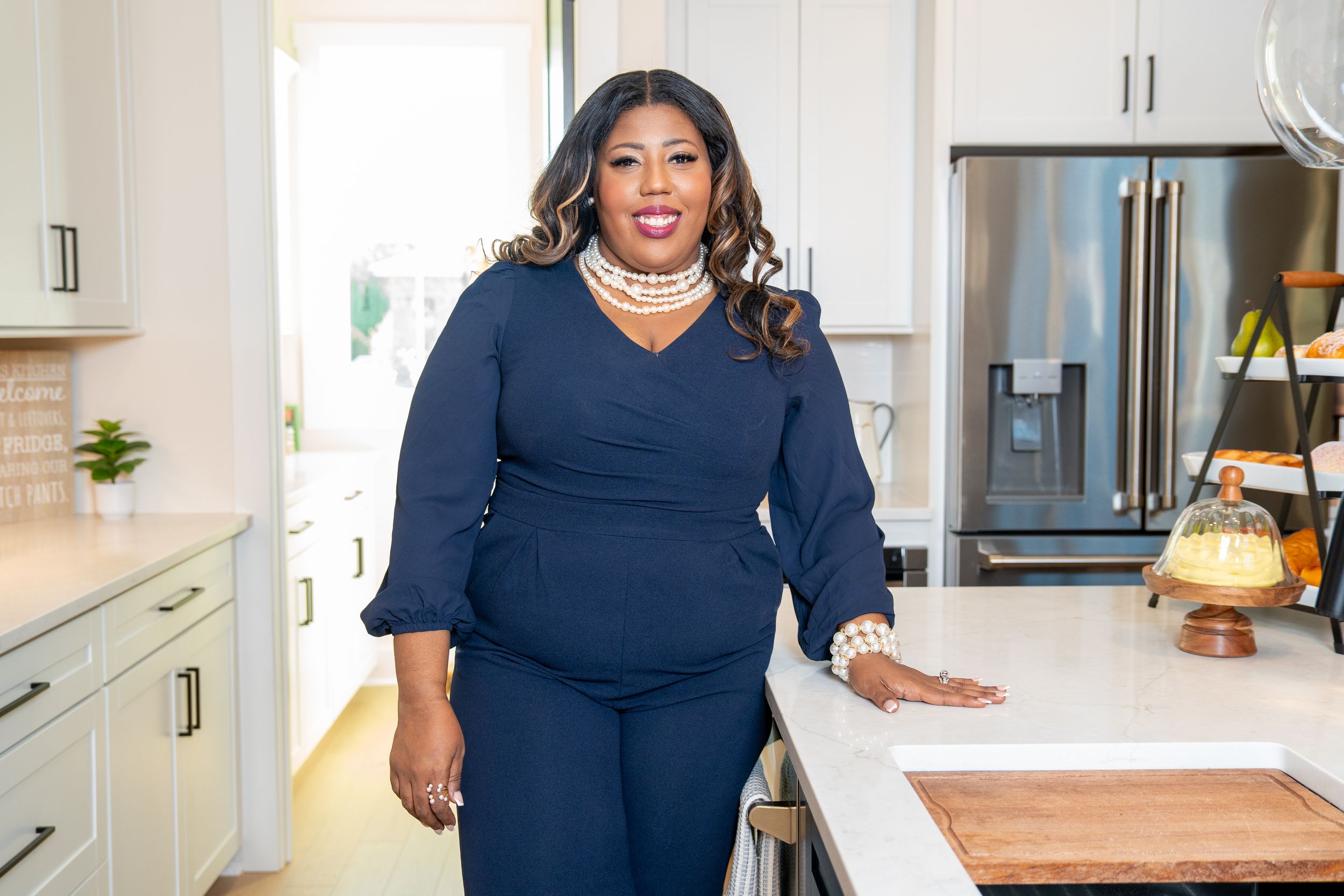 A confident woman in a navy blue outfit and layered pearl necklace leans on a white kitchen island in a bright home.