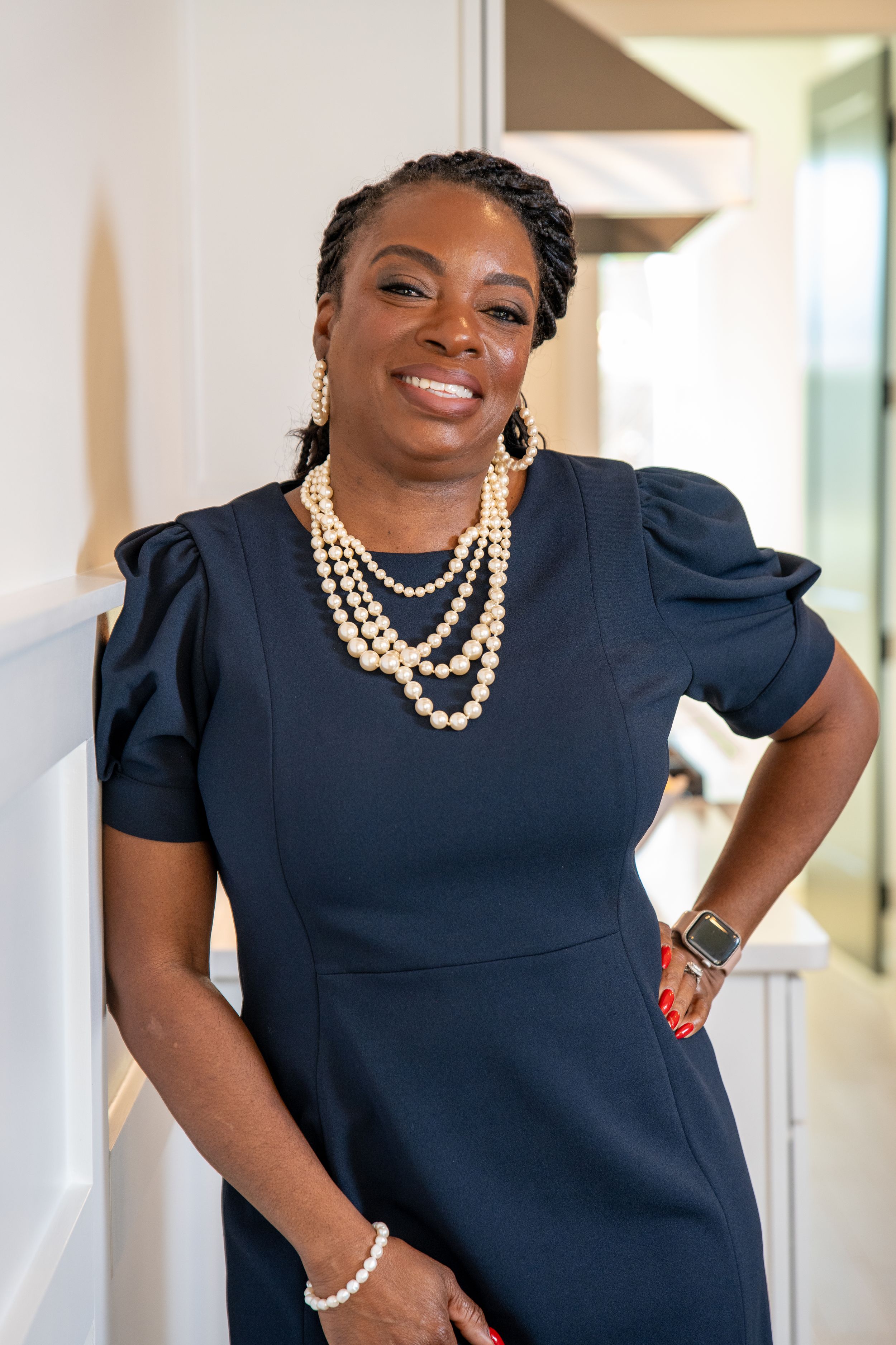 A smiling woman in a navy dress with puff sleeves, a pearl necklace and a smartwatch poses with a hand on her hip.