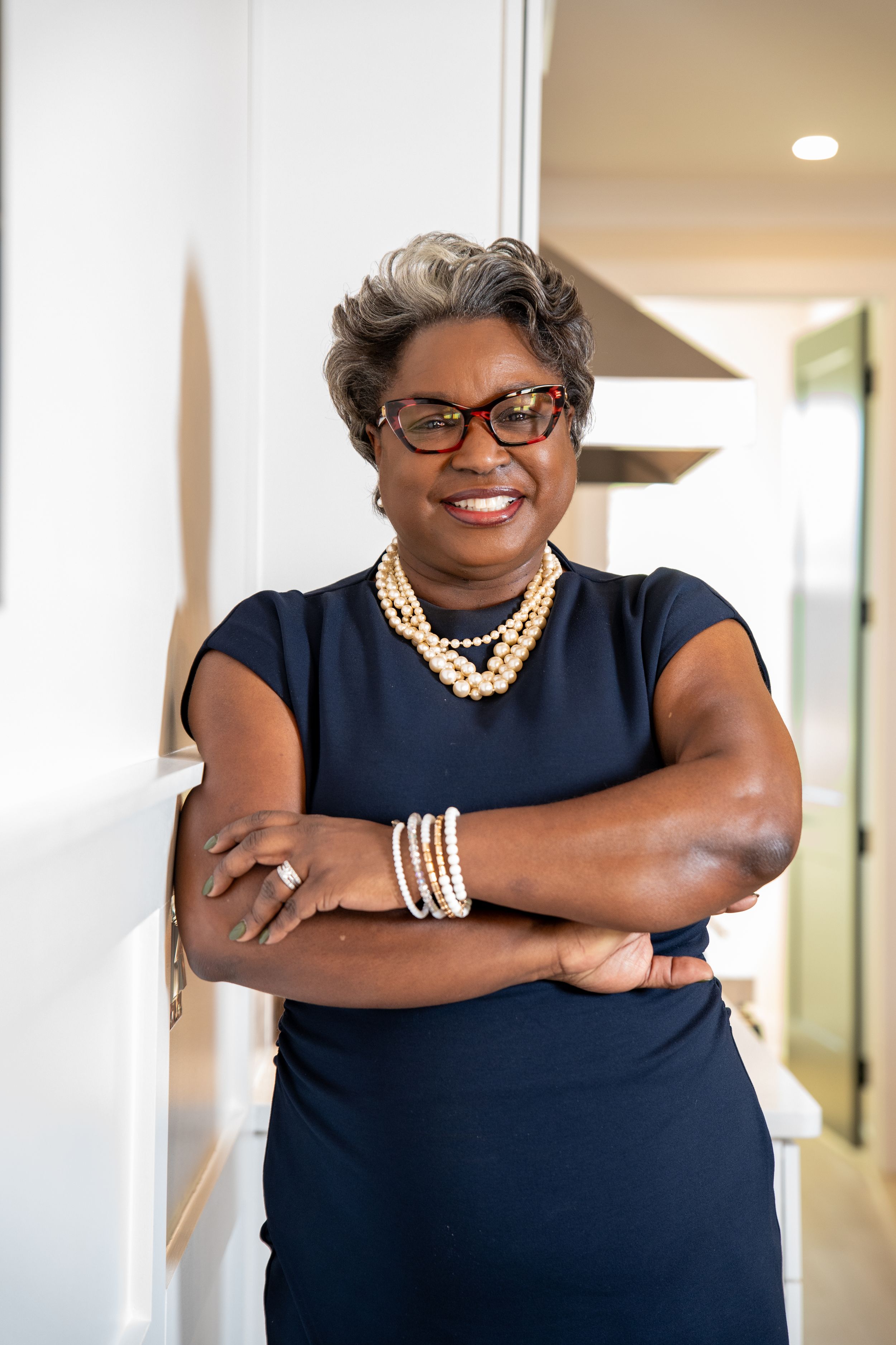 A woman with short gray hair, red glasses and a pearl necklace smiles with her arms crossed in a bright, modern home.