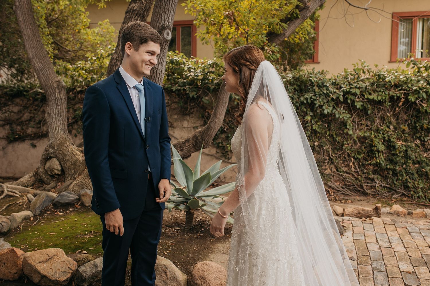 Bride in lace gown and veil smiles with groom in navy suit outdoors at Rancho Buena Vista Adobe.