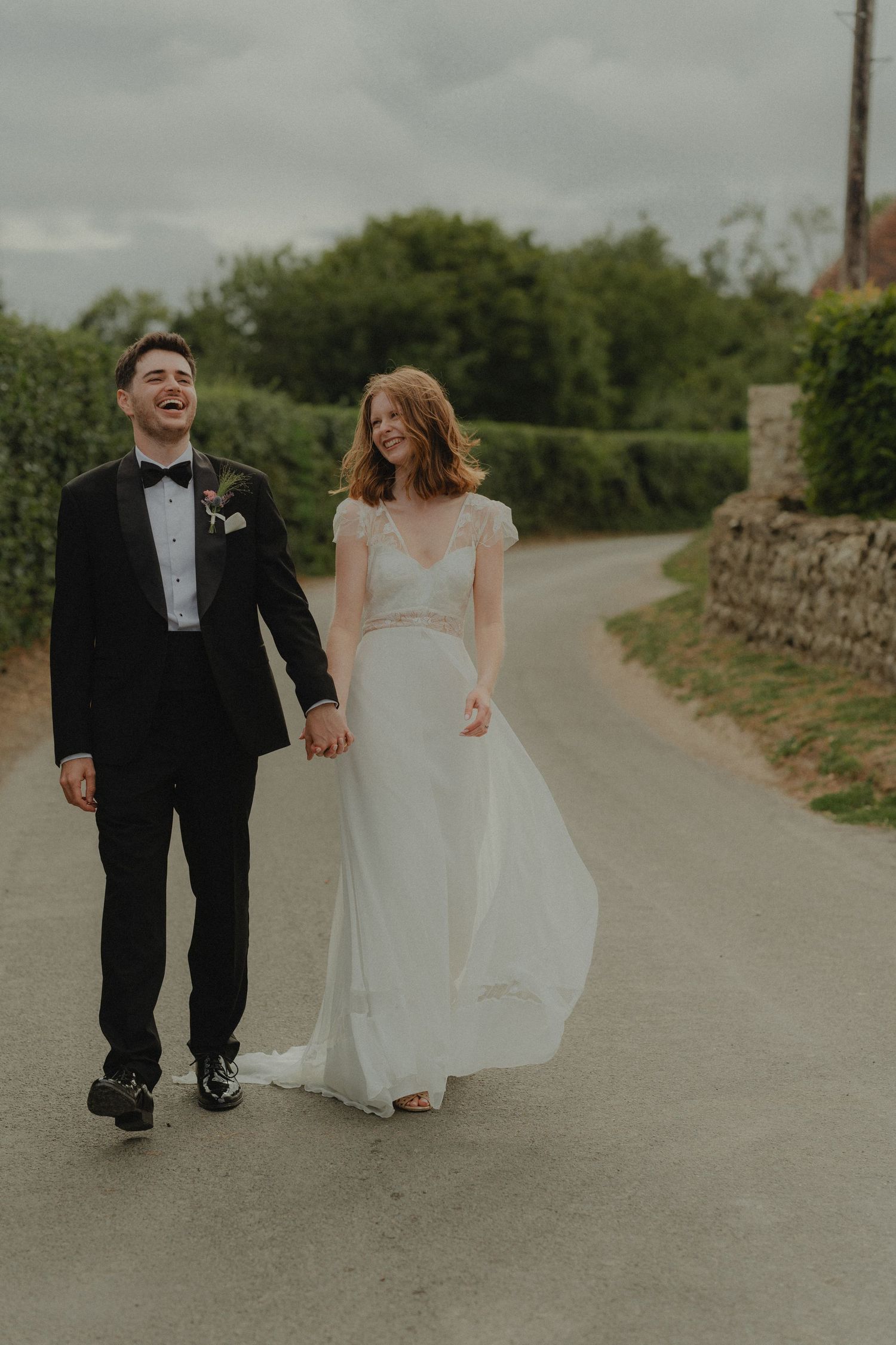 A laughing bride and groom walk hand in hand down a country road, she in a flowing white gown, he in a black tuxedo.