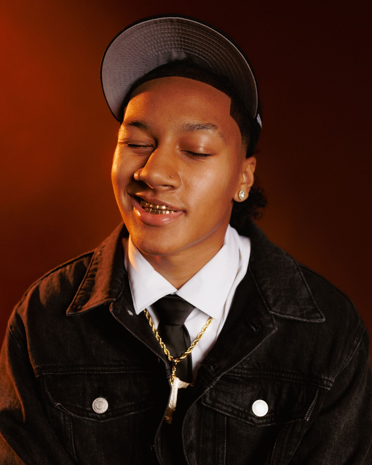 A person in a black jacket and white shirt smiles with closed eyes against a dark brown background.