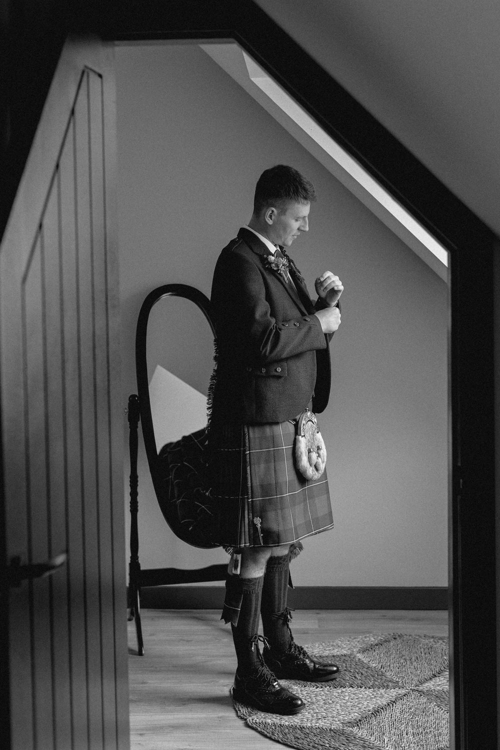 Black and white photo of someone in traditional Scottish kilt and formal attire adjusting their outfit in a hallway.