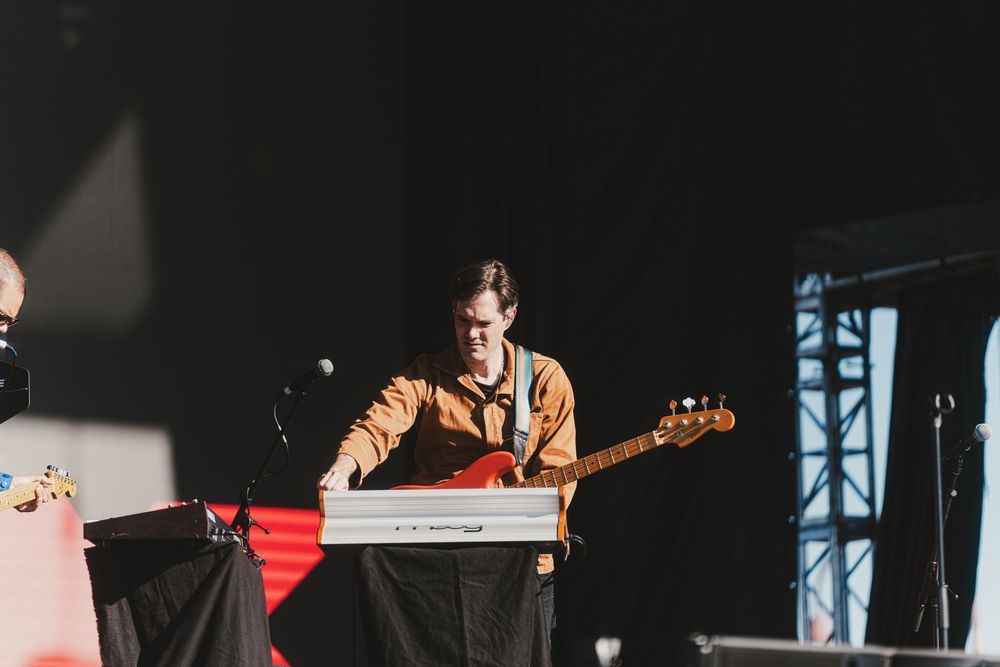 Musicians perform on stage at an outdoor concert with lighting and sound equipment visible in the background.