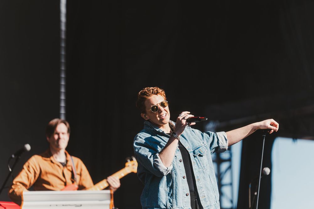 Performers on stage during a live music show with one singing into a microphone while another plays instruments.