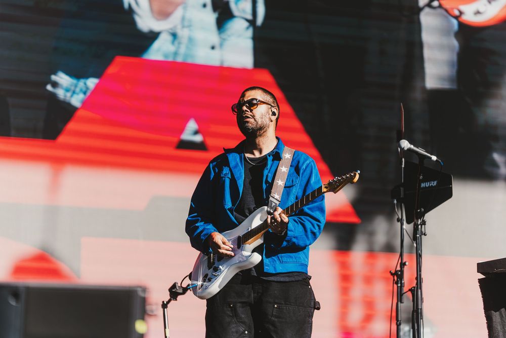 A musician in a blue shirt plays guitar on stage with dramatic red lighting in the background.