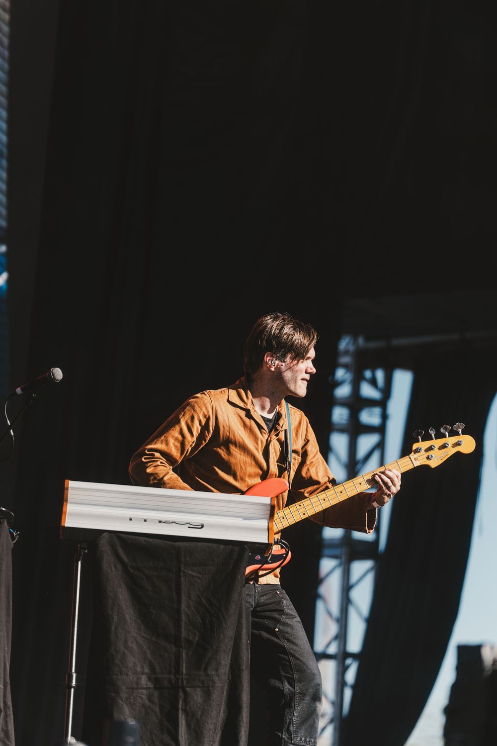 Musician in brown jacket performs on stage with guitar during outdoor concert performance.