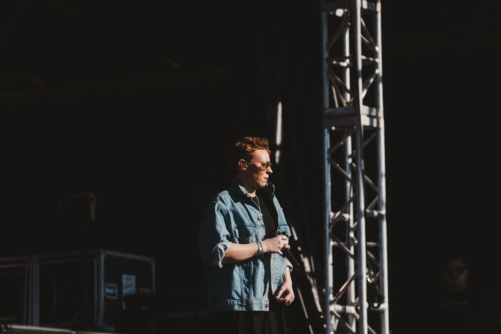 A performer in denim stands on stage with dramatic lighting and metal scaffolding structures visible in the background.
