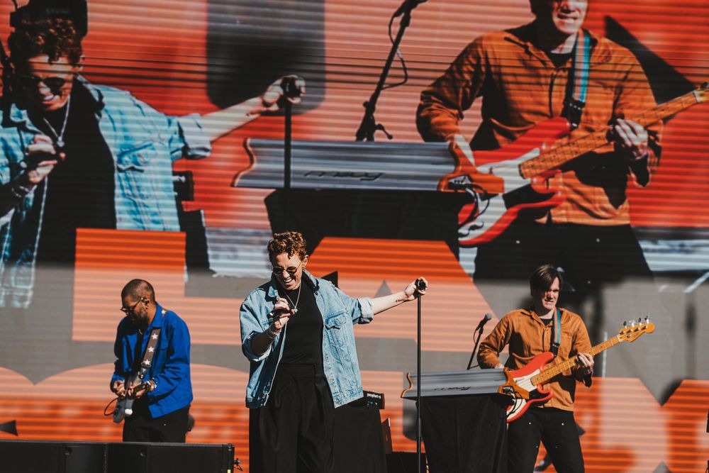 Musicians perform on stage with guitars and instruments against a vibrant orange and blue backdrop during a live concert.