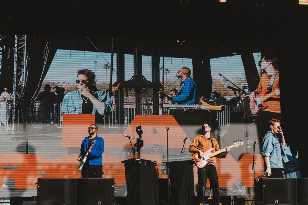 Musicians perform on stage at an outdoor concert venue with orange lighting and dark background.