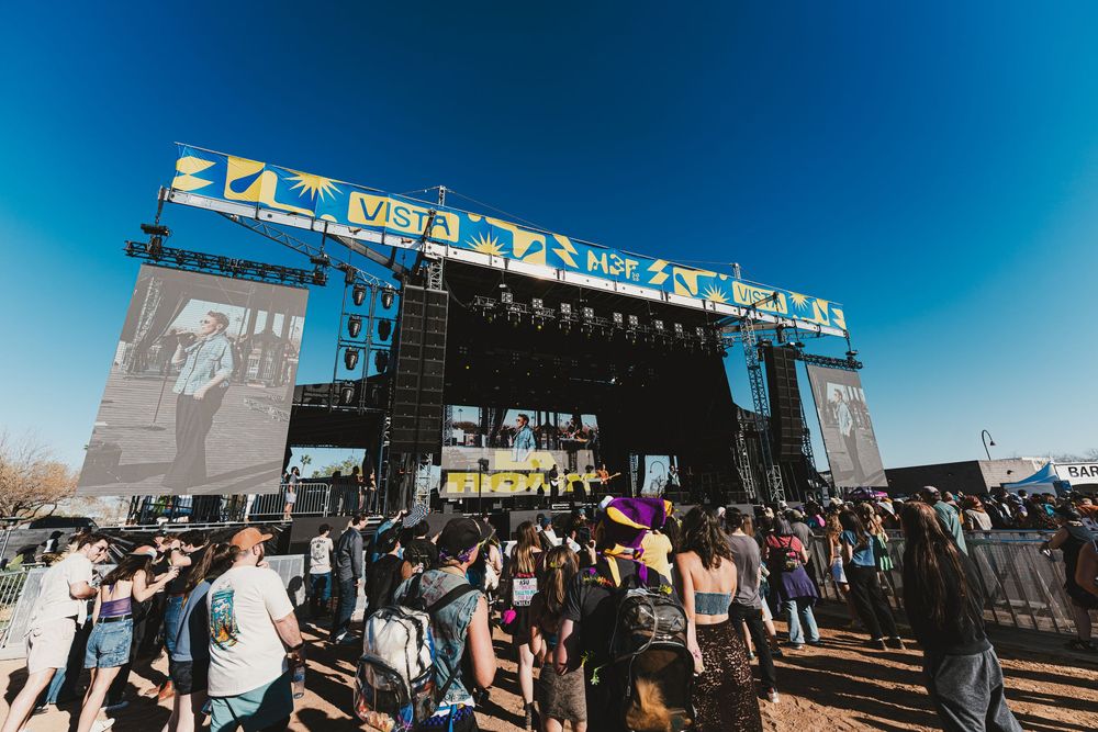 Music festival crowd gathers in front of large outdoor stage during daytime performance.