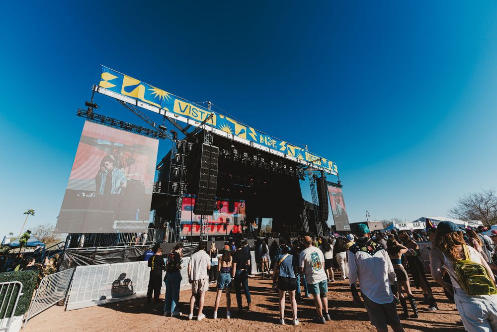 Panoramic view of a festival entrance with crowds gathering outside under bright blue sky and white signage.