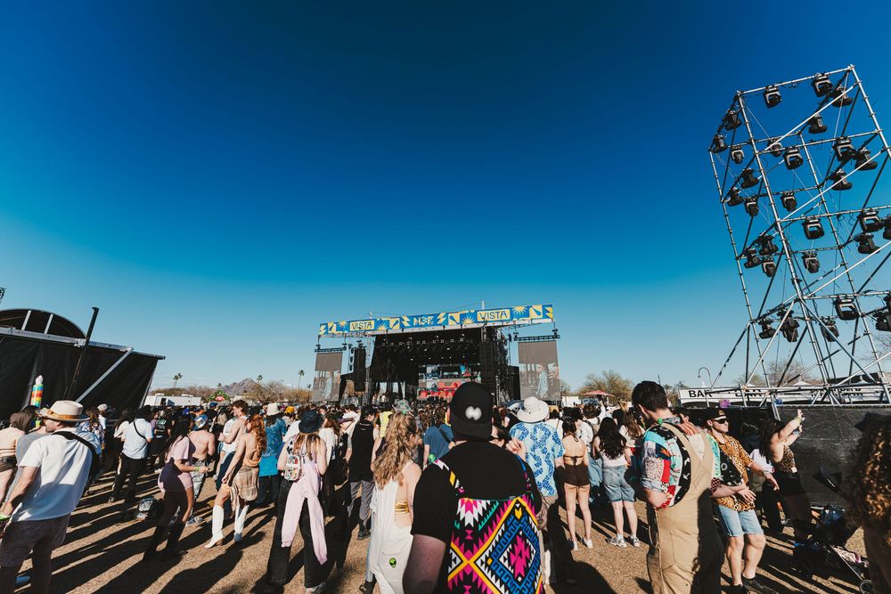 Outdoor music festival crowd gathered in front of a stage under bright blue sky with metal scaffolding visible.