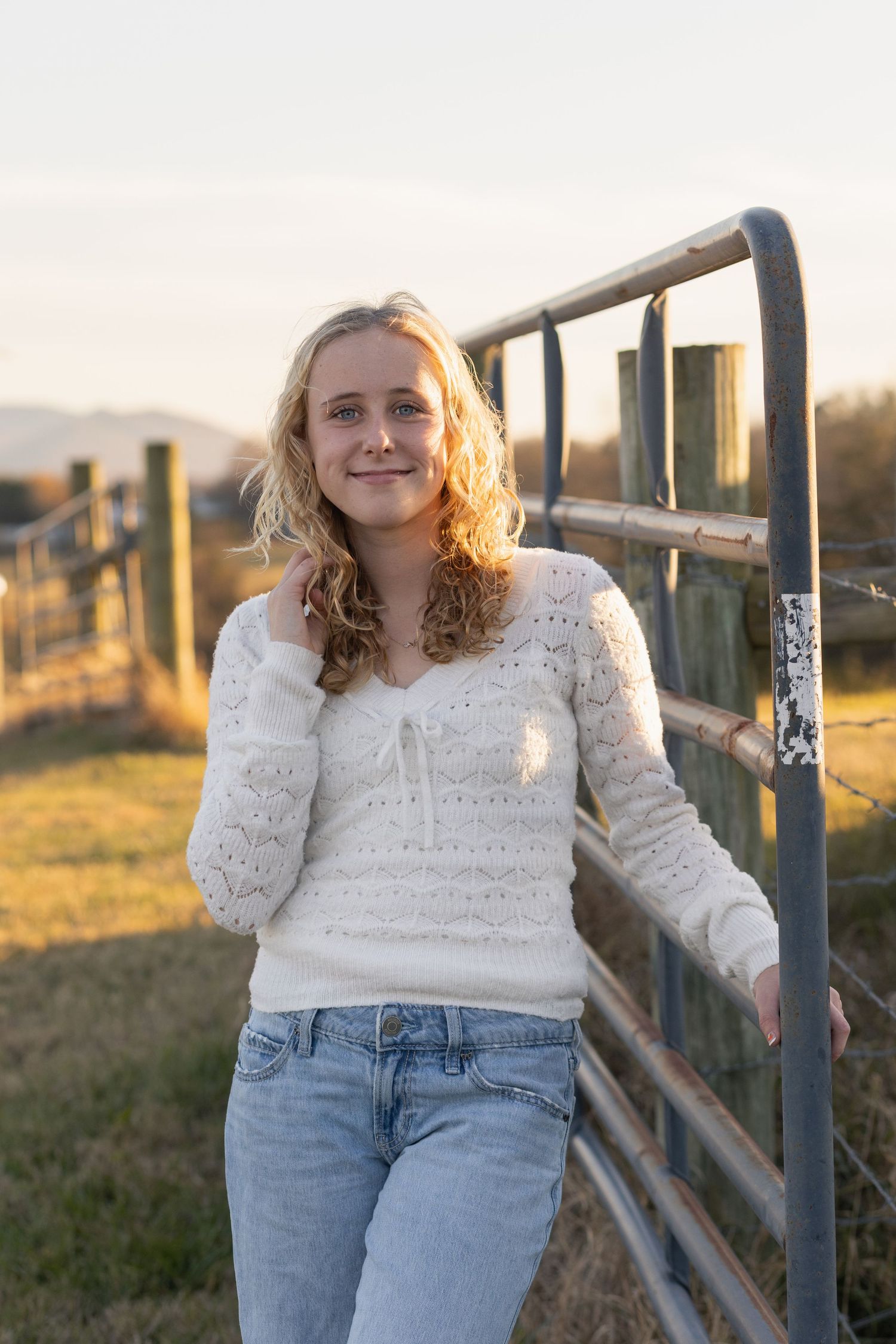 A woman in a white lace sweater and jeans leans against a wooden fence at sunset in a pastoral setting.