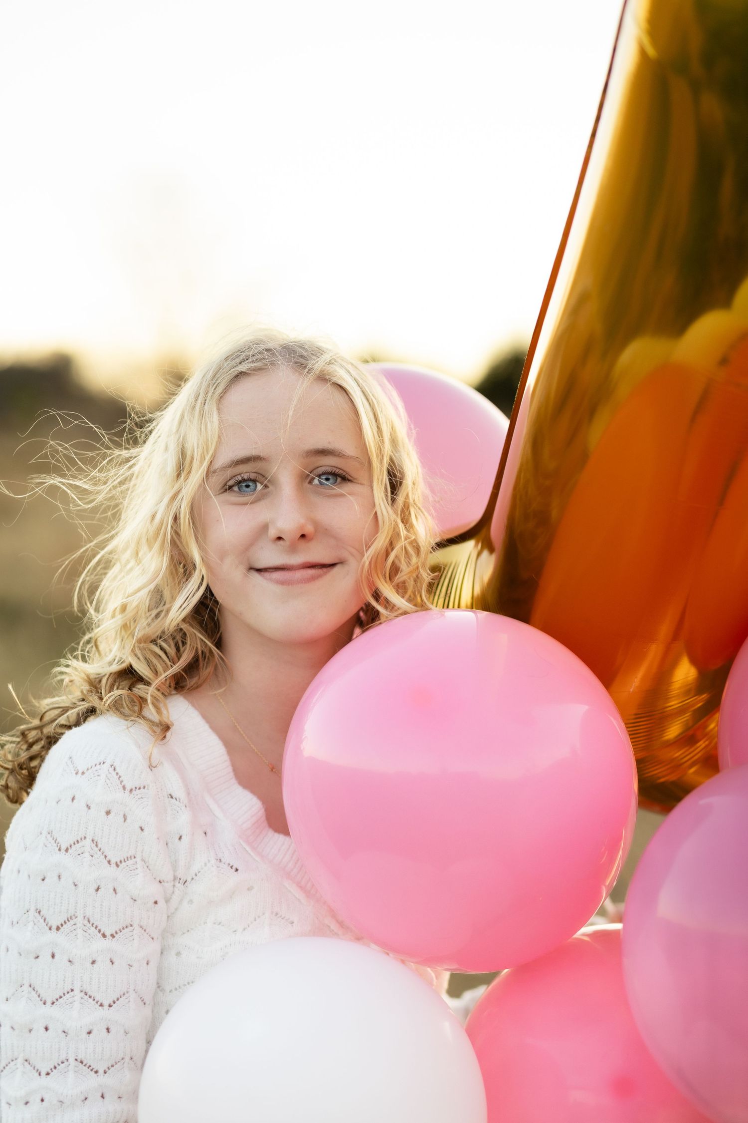 A person in a white lace top poses with pink and white balloons against a warm sunset background.
