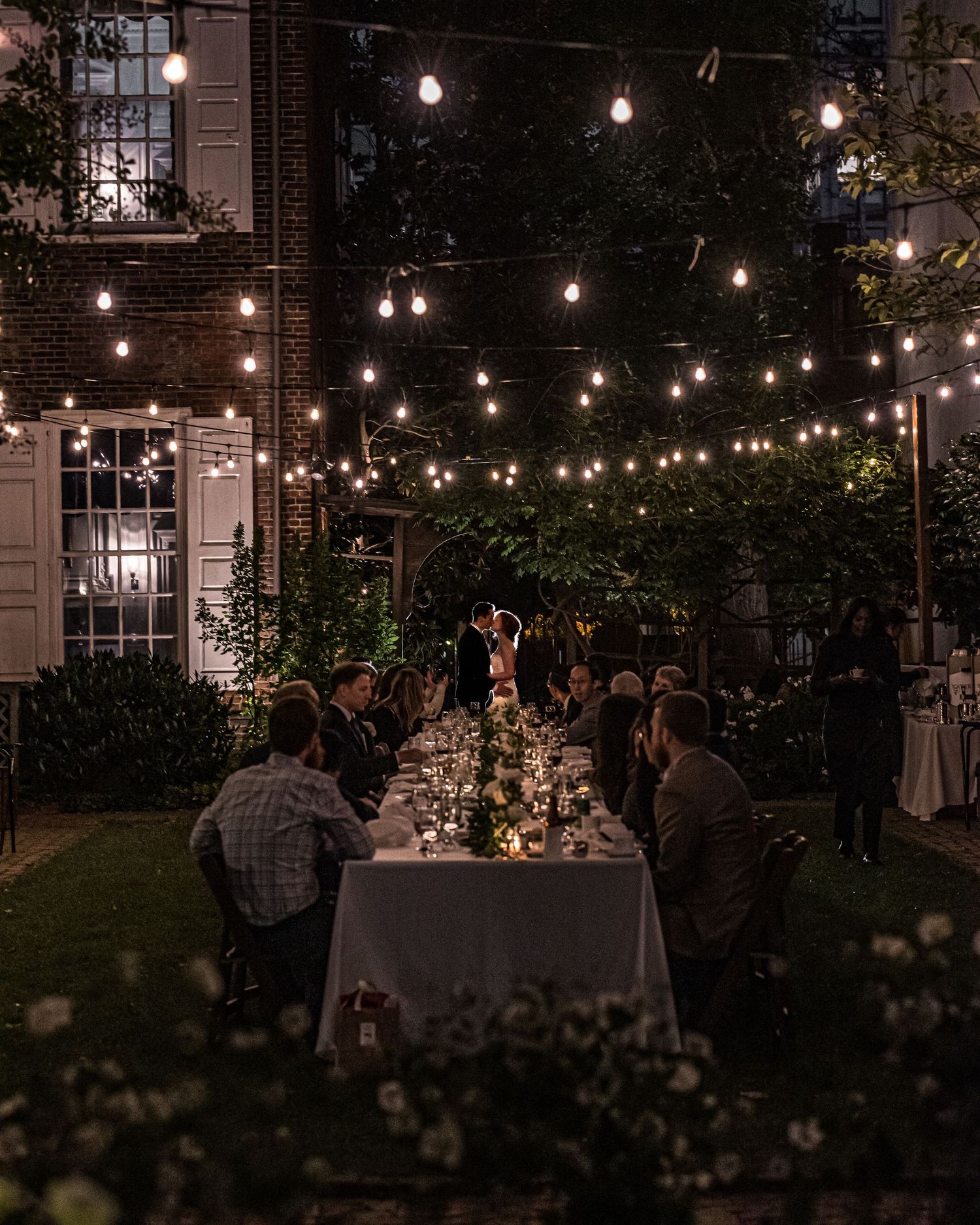 String lights illuminate an outdoor evening garden party with guests dining at tables under a dark sky.