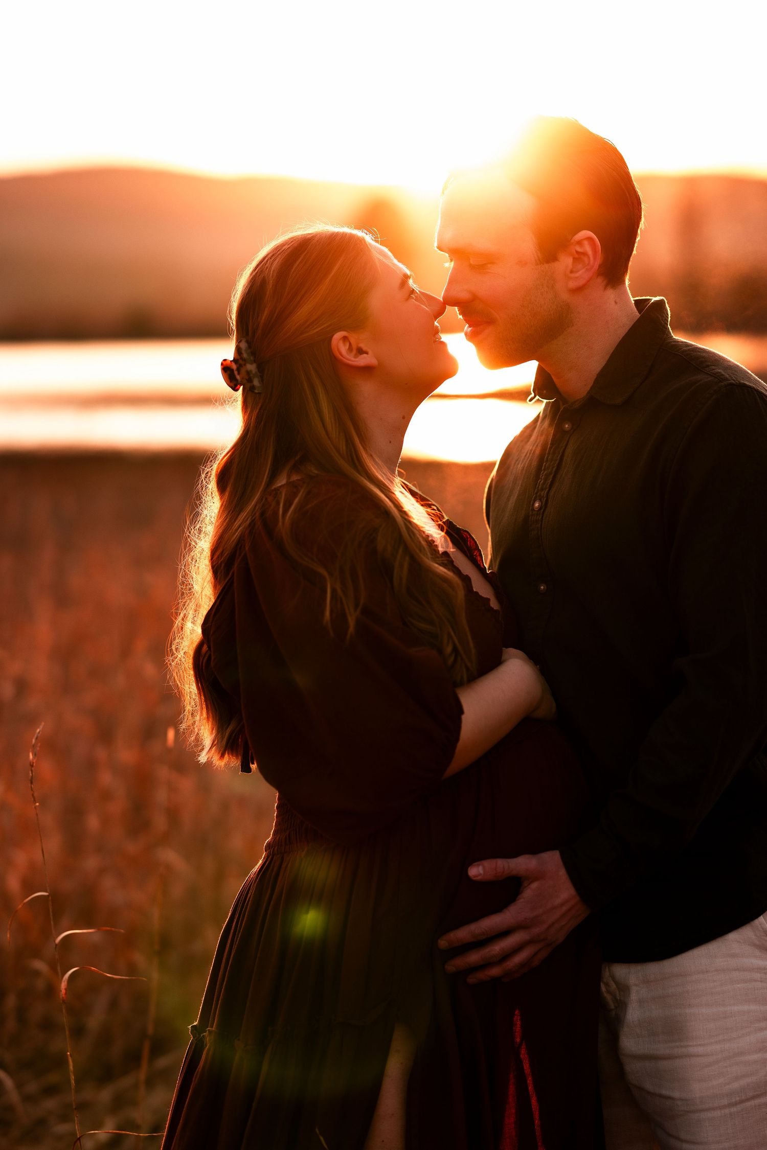 A romantic couple embraces in a golden sunset field with warm backlighting creating a dreamy silhouette effect.