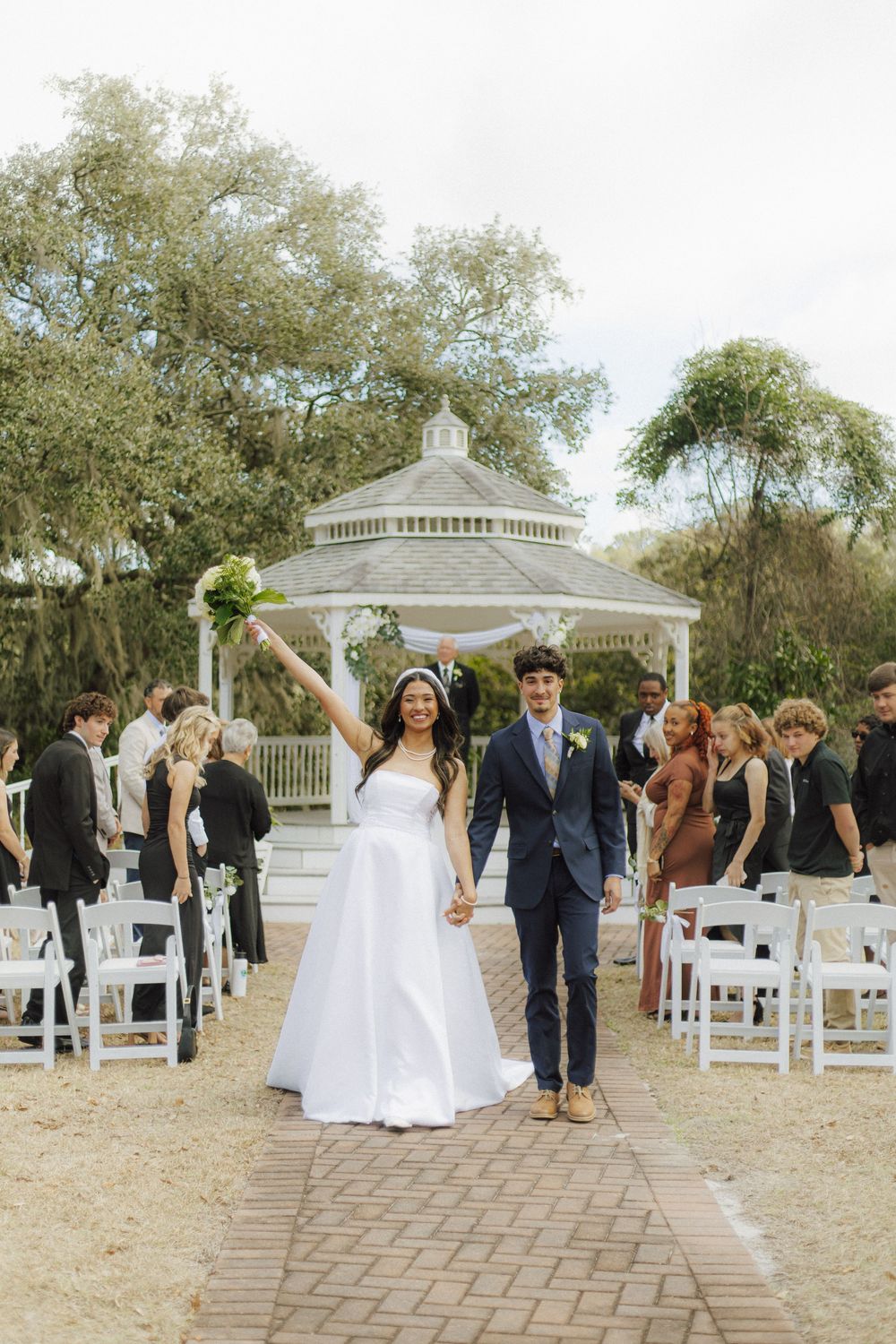 Wedding guests celebrate at an outdoor ceremony with a white gazebo and chairs on brick pavers.