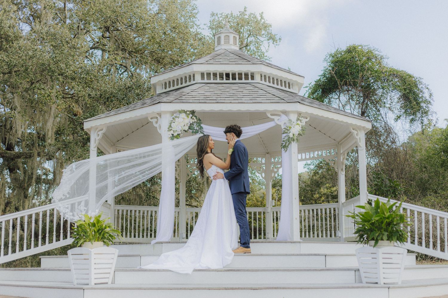 A white wedding gazebo surrounded by lush trees and landscaping in a romantic outdoor setting.