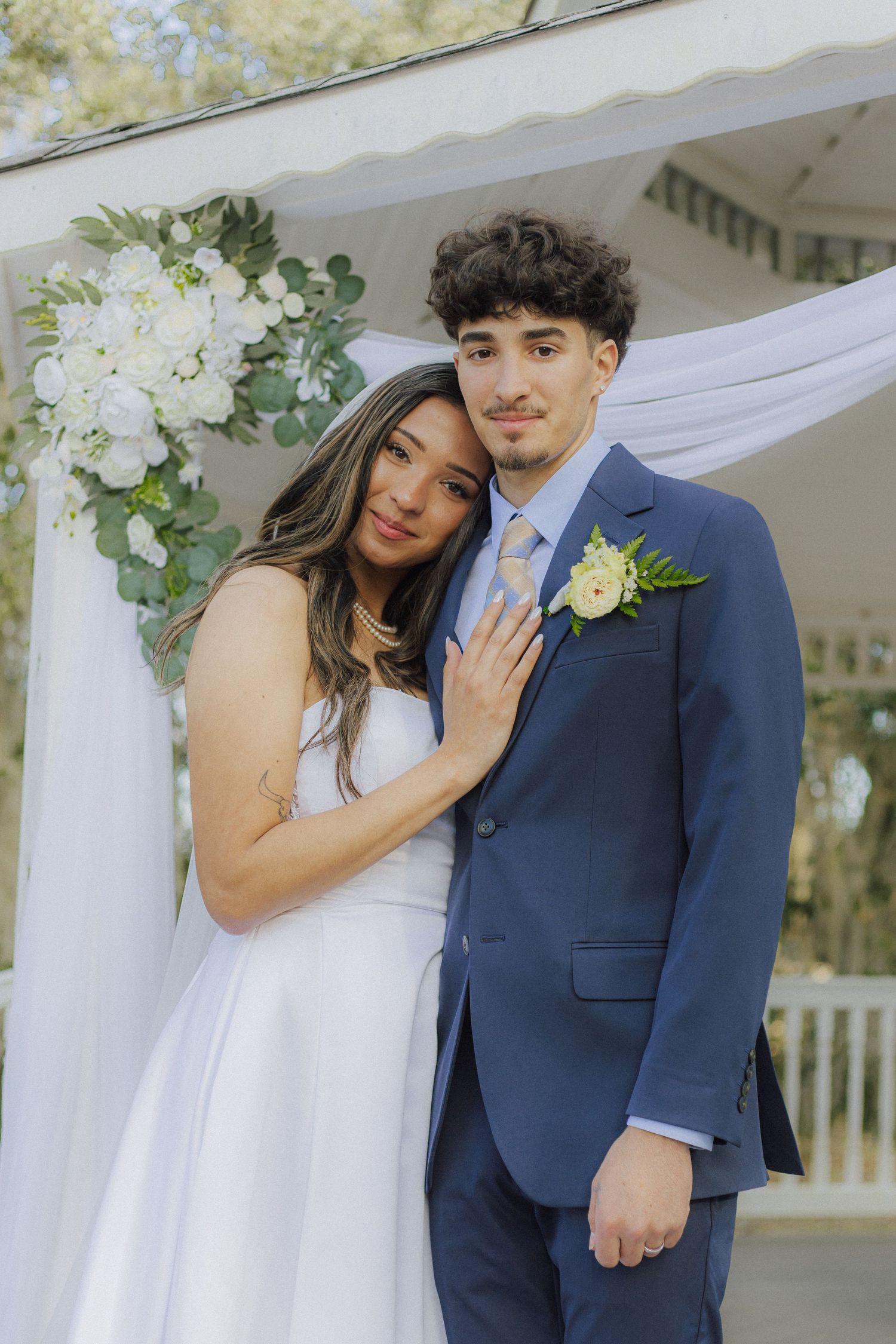 Bride and groom pose under floral gazebo, she in white dress, he in navy suit with boutonniere.