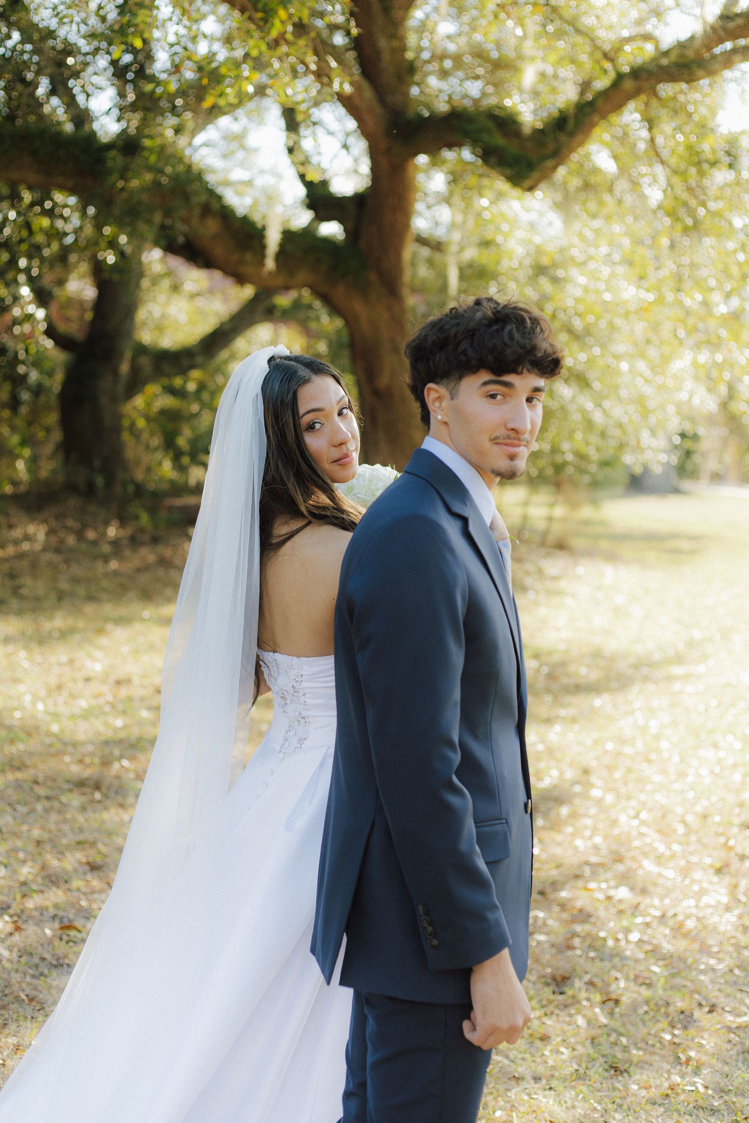 A bride in white gown and groom in navy suit pose together beneath sprawling tree branches in golden sunlight.