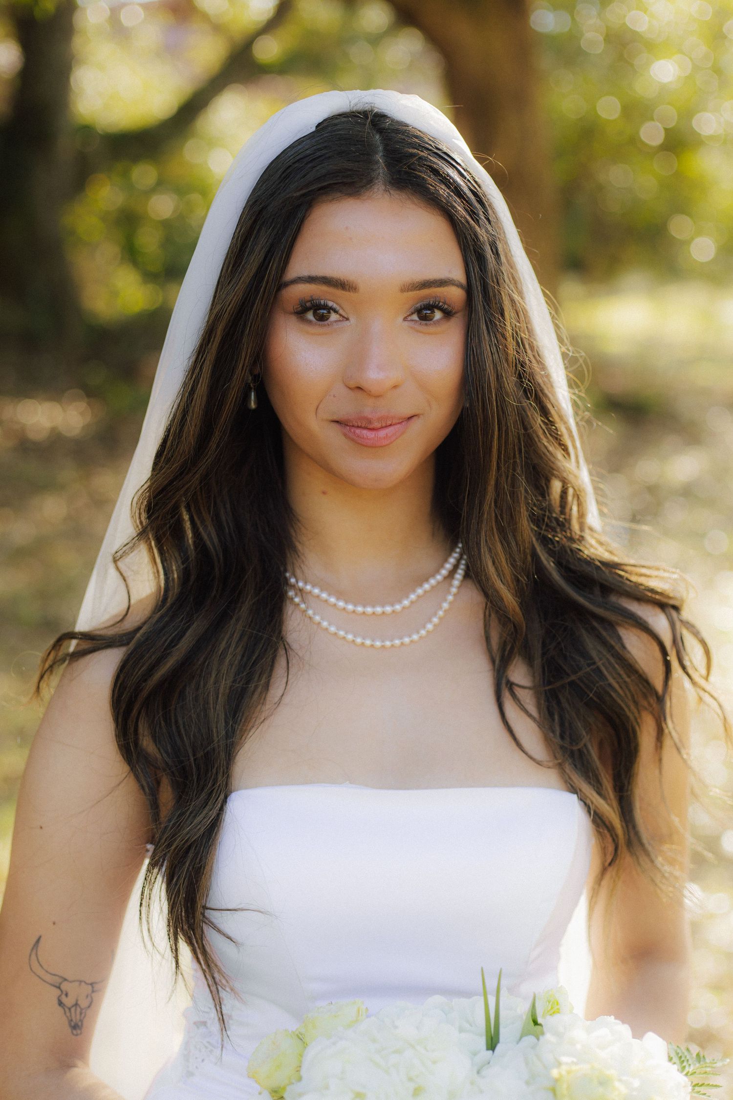 Bride in white strapless gown and veil with pearl necklace holding white bouquet outdoors.