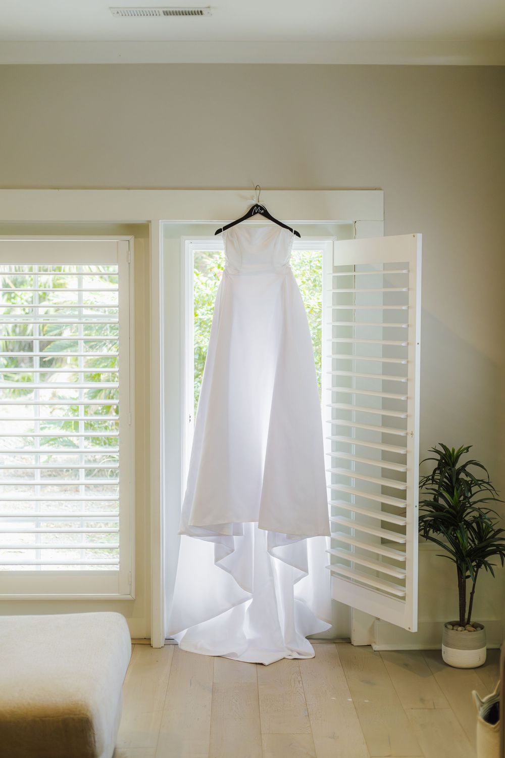 A white wedding dress hangs elegantly on a hanger against white plantation shutters with natural light streaming through.