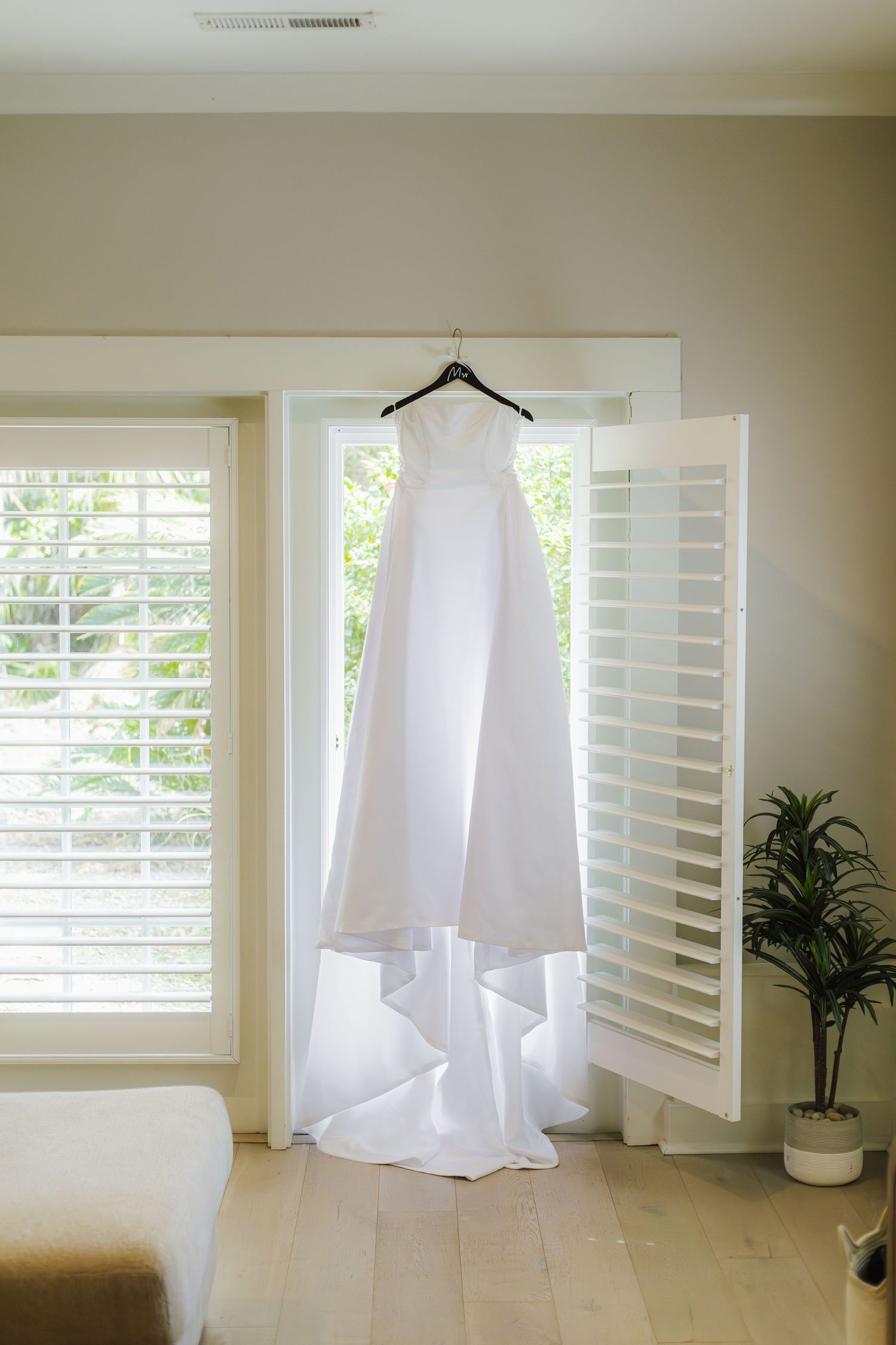 A white wedding dress hangs elegantly on a hanger against white plantation shutters with natural light streaming through.