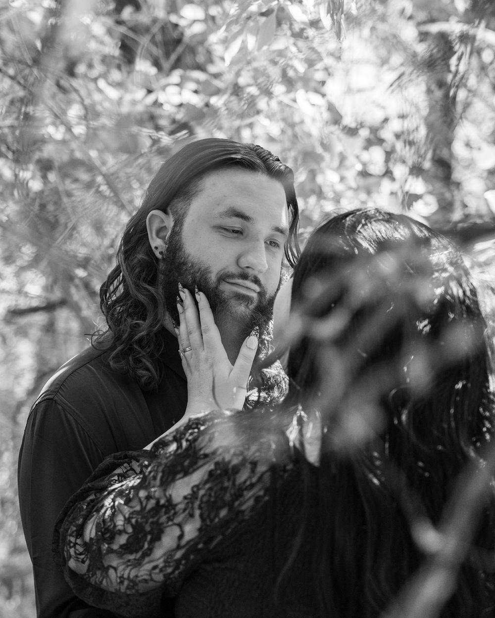 Black and white romantic photo of a bearded man with long hair and a woman in lace, sharing an intimate moment outdoors.