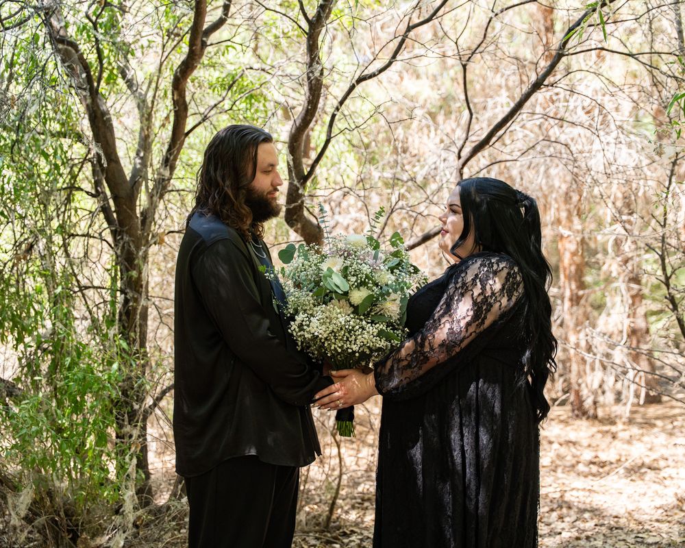 A couple in black gothic attire exchanges bouquet of white flowers in a woodland setting.