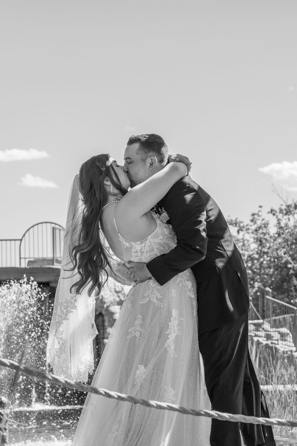 Black and white photo of a bride and groom sharing a romantic kiss near a fountain on their wedding day.