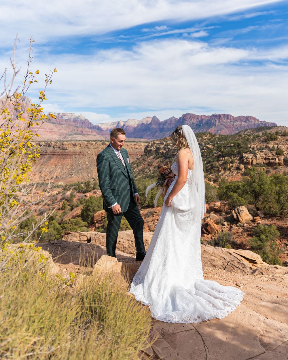 A couple in wedding attire poses on red rock formations with desert mountains and blue skies in the background.